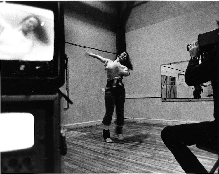 b and wphoto , white female with long dark hair, in the corner of a studio with white walls, one mirrored wall, is being filmed by a man who we partially see, wearing black, to his left are video monitors and we can see the woman's face reflected in top monitor